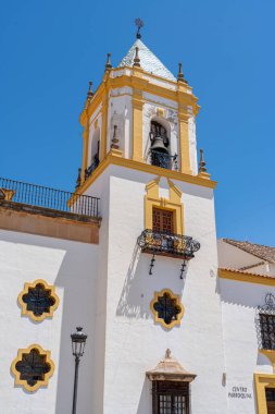 İspanya 'nın Ronda kentindeki tarihi bir kilise çan kulesinin yakından görüntüsü, açık mavi gökyüzüne karşı klasik sarı ve beyaz mimari detaylar içeriyor. Kültürel ve mimari temalar için ideal.