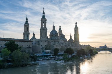 İspanya 'nın Zaragoza kentindeki Bazilika del Pilar' da gün batımında, Ebro Nehri 'nin huzurlu yansımalarıyla aydınlandı. Seyahat, mimari ve kültürel temalar için ideal.
