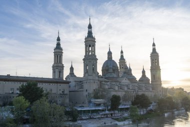 İspanya 'nın Zaragoza kentindeki Bazilika del Pilar' da gün batımında, Ebro Nehri 'nin huzurlu yansımalarıyla aydınlandı. Seyahat, mimari ve kültürel temalar için ideal.