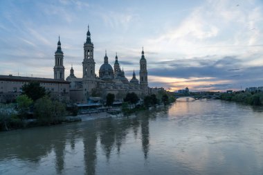 İspanya 'nın Zaragoza kentindeki Bazilika del Pilar' da gün batımında, Ebro Nehri 'nin huzurlu yansımalarıyla aydınlandı. Seyahat, mimari ve kültürel temalar için ideal.