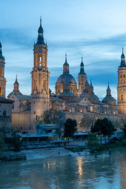 İspanya 'nın Zaragoza kentindeki Bazilika del Pilar' da gün batımında, Ebro Nehri 'nin huzurlu yansımalarıyla aydınlandı. Seyahat, mimari ve kültürel temalar için ideal.
