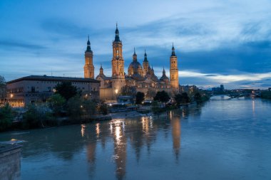 İspanya 'nın Zaragoza kentindeki Bazilika del Pilar' da gün batımında, Ebro Nehri 'nin huzurlu yansımalarıyla aydınlandı. Seyahat, mimari ve kültürel temalar için ideal.