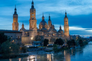 İspanya 'nın Zaragoza kentindeki Bazilika del Pilar' da gün batımında, Ebro Nehri 'nin huzurlu yansımalarıyla aydınlandı. Seyahat, mimari ve kültürel temalar için ideal.