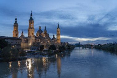 İspanya 'nın Zaragoza kentindeki Bazilika del Pilar' da gün batımında, Ebro Nehri 'nin huzurlu yansımalarıyla aydınlandı. Seyahat, mimari ve kültürel temalar için ideal.