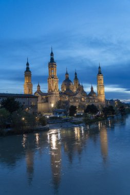 İspanya 'nın Zaragoza kentindeki Bazilika del Pilar' da gün batımında, Ebro Nehri 'nin huzurlu yansımalarıyla aydınlandı. Seyahat, mimari ve kültürel temalar için ideal.