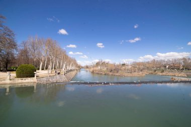 Resimli bir bahar gününün tadını çıkaran ziyaretçiler İspanya 'nın Aranjuez kentindeki nehir kıyısında geziniyorlar. Sahne canlı yeşillik ve parlak mavi gökyüzü ile güçlendirilir..