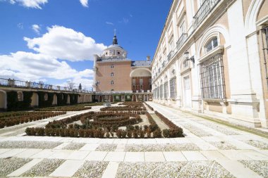 Ziyaretçiler, Aranjuez 'deki kraliyet sarayının etrafındaki titizlikle korunan bahçeyi keşfediyorlar. Parlak mavi gökyüzü zarif mimari ve yeşilliği tamamlıyor..