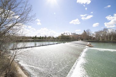 Aranjuez, İspanya 'da sakin bir turkuaz göle akan sakin bir şelale. Etrafı ağaçlarla çevrili ve mavi gökyüzü altında tüylü bulutlarla kaplı tarihi binalar..