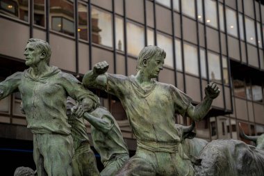 San Fermin festivalinde boğaların ünlü koşusunu anan bronz bir heykel Pamplona, Navarra 'da duruyor. Çevredeki binaların yansımaları olay yerine derinlik katıyor..