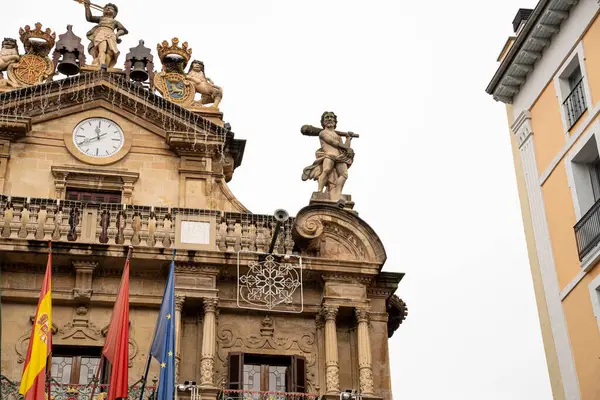 Pamplona 'daki etkileyici belediye binası süs, saat ve mermer heykelini gri gökyüzüne karşı sergiliyor. Rüzgarda dalgalanan bayraklar şehrin zengin kültürünü ve tarihini vurguluyor..