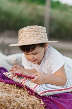 Little boy in straw hat sitting on the pebbles and looking at the phone. High quality photo