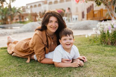 Parktaki çimlerin üzerinde oturan ve kameraya gülümseyen oğluyla mutlu bir anne. Ebeveynlik, aile, çocuk gelişimi ve doğada eğlence. Yüksek kalite fotoğraf