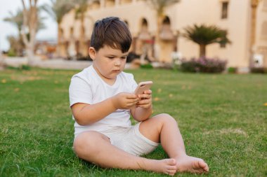 Sarışın, kıvırcık saçlı, açık yeşil çimenlerin üzerinde oturan, açık havada güneşli bir günde elinde cep telefonu olan, küçük, sevimli, meraklı bir oğlan çocuğu. Yüksek kalite fotoğraf