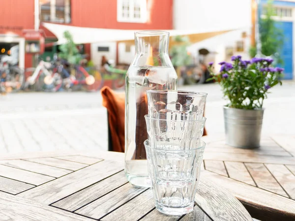 Three glasses and a carafe with water and ice cubes at an outdoor restaurant table. Shallow depth of field with glasses and carafe in focus. Flowers and bicycles out of focus in the background.