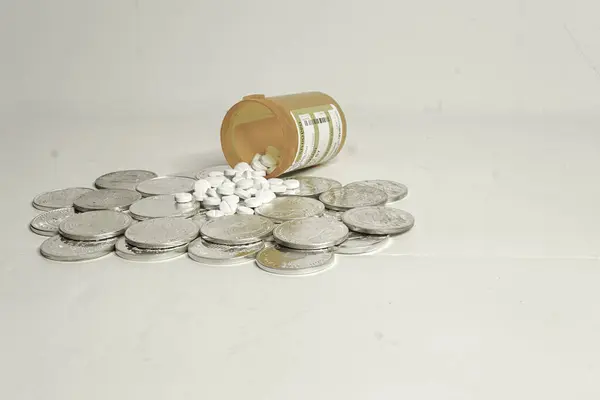 Bir reçete şişesi yan yatmış bir yığın gümüş doların üzerine tabletleri döküyor. Konsept, reçetelerin masraflarıdır..