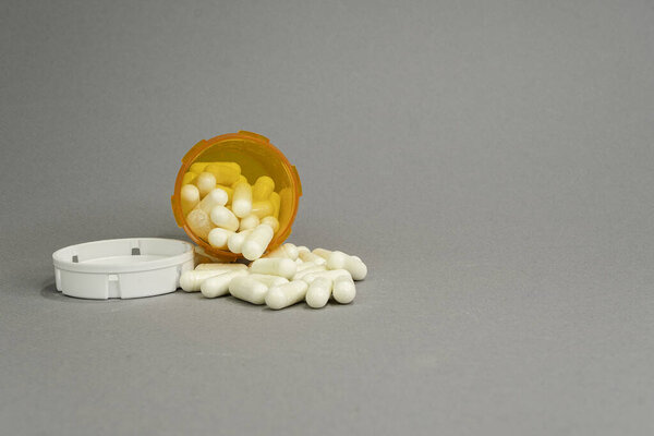 Открытая оранжевая бутылка с рецептом на боковом фронте, которая имеет несколько капсул, выплеснутых на сером фоне.