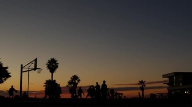 Basketbol oynayan insanlar, basketbol sahasındaki oyuncuların siluetleri, günbatımı okyanus sahili, California sahili, Mission plajı, ABD. Sokak topu sahasında siyah halka, ağ ve arkabahta.