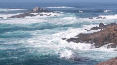 Kayalık okyanus plajı. Büyük dalgalar kayalıklara çarpıyor, mavi su sıçrıyor, deniz köpüğü. Big Sur yakınlarında doğanın gücü, 17 millik yol. Dramatik deniz manzarası. Point Lobos, Monterey, California sahili, ABD