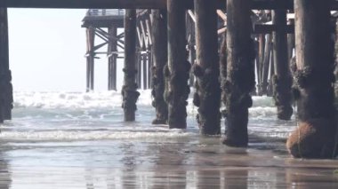 Kümelerdeki ahşap kristal iskelenin altında, Kaliforniya 'daki okyanus sahili su dalgaları. San Diego sahilinde yaz tatili. Deniz kıyısındaki rıhtımın altında. Kusursuz döngülü görüntü grafiği.