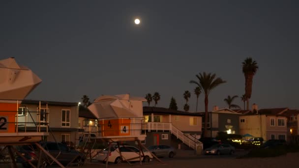 ヤシの木のシルエットと夕暮れの空の満月 カリフォルニアビーチ 夕方には海岸沿いの海辺の家や家 夕暮れ時には太平洋岸の満月 ライフガードタワー 窓の光 — ストック動画