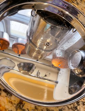 A colorful alternative view inside a kichen with brightly polished pots, pans, and other cooking items hanging from the ceiling, reflecting in a pot lid resting on a marble counter top. 