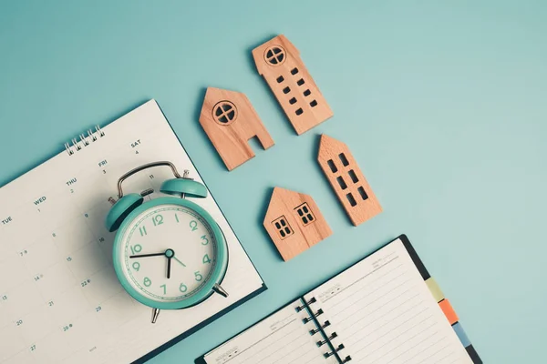 An alarm clock with calendar, notebook and wooden house model for the concept of time management, housing and property.