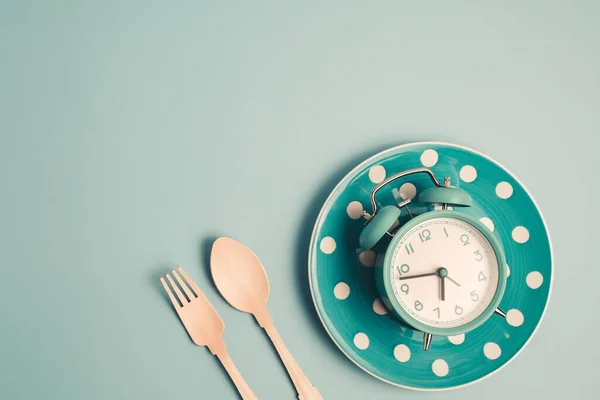 An alarm clock on an empty plate and cutlery set against blue background for the concept of food, time management, losing weight and eating on time.