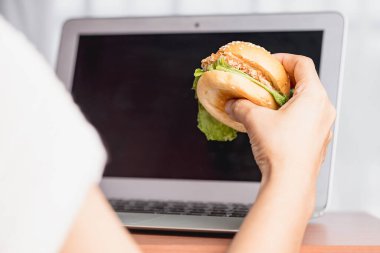 Lezzetli bir hamburgeri elle tutmak ve çalışırken yemek için dizüstü bilgisayar kullanmak..