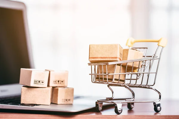 Brown parcel boxes on the laptop and in the shopping cart are positioned on a wooden table for the concept of online shopping, convenience and purchases arriving at doorstep.