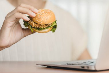 Peynirli, marullu, domatesli ve biftekli lezzetli bir hamburger yemeğinin tadını çıkaran bir insan, iş yerinde fast food yemek için ellerinde tutulur.