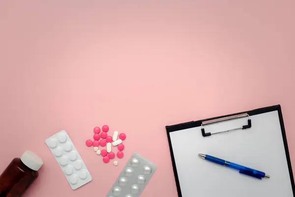 Flat lay of a bottle of medicine, medication blister packs, pills, clipboard and pen on a pink background for the concept of medical note taking and healthcare