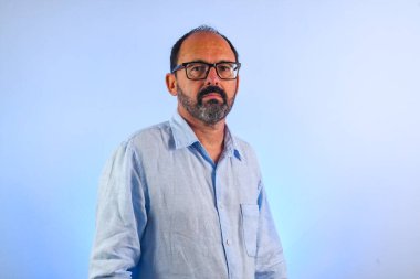 60 yaşında bir adam mavi arka planda kameraya bakıyor.