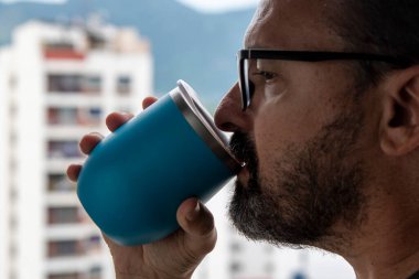 Bulanık şehir arka planına bakan mavi bardaktan su içen adam.