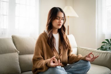 Genç Asyalı kadın meditasyon yapıyor ve evdeki oturma odasında oturuyor.