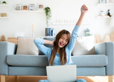Genç Asyalı kadın evde dizüstü bilgisayarla çalışırken kanepenin yanında oturuyor. Heyecanlı bayan kazananımız, başarıyı kutlarken bir yandan da deftere bakıyor.