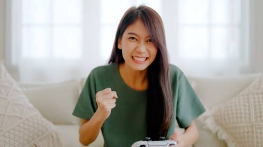 Beautiful young asian woman sitting on sofa and holding joystick playing online video game in living room at house