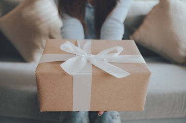 Genç Asyalı bir kadın hediye kutusu tutuyor ve oturma odasında kanepede oturuyor. Mutlu yıllar, Şükran Günü, Mutlu Noeller, Mutlu yıllar