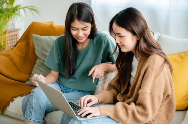 İki Asyalı genç kadın mutlu bir şekilde gülümsüyor ve bilgisayarını evdeki oturma odasındaki koltukta kullanıyor.