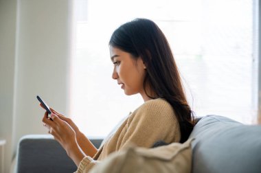 Happy young asian woman relax on comfortable couch at home texting messaging on smartphone, smiling girl use cellphone chatting, browse wireless internet on gadget, shopping online from home