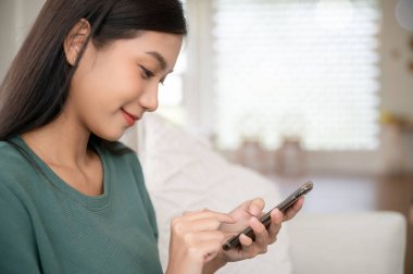 Görüntüyü kapat. Mutlu Asyalı kadın evinde dinleniyor. Kanepede oturuyor ve cep telefonu kullanıyor.