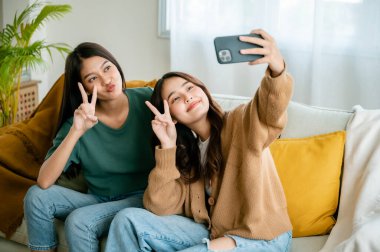 İki Asyalı genç kadın mutlu mesut gülümsüyor ve evdeki oturma odasında selfie çekiyor.