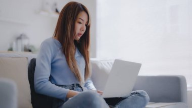 Genç Asyalı kadın evdeki koltukta otururken dizüstü bilgisayar kullanıyor.