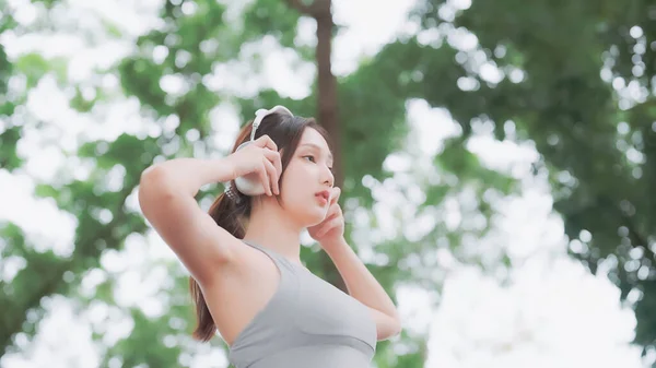 Asian woman trainer is engaged in fitness in public park. Female wearing sport bra and using headphone listening to music. Healthy lifestyle, sports outdoor activities in park