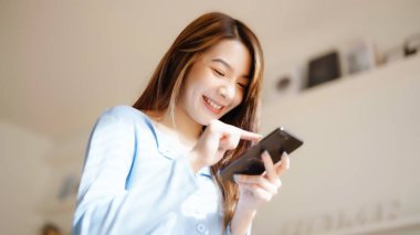 Mutlu genç Asyalı kadın rahat bir kanepede rahat rahat oturup akıllı telefonuna mesaj atıyor, gülümseyen kız cep telefonu sohbetini kullanıyor, cihazda kablosuz interneti tarıyor, evden online alışveriş yapıyor.