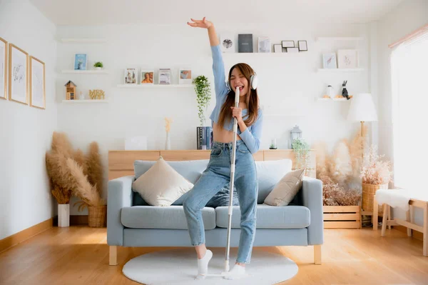House cleaning with fun. Happy young asian housewife singing her favorite song during cleanup, using mop as microphone, enjoying domestic work. Young woman dancing and cleaning in living room