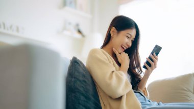 Happy young asian woman relax on comfortable couch at home texting messaging on smartphone, smiling girl use cellphone, chatting online message, shopping online from home