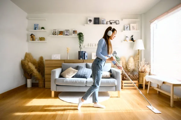 House cleaning with fun. Happy young asian housewife singing her favorite song during cleanup, using mop as microphone, enjoying domestic work. Young woman dancing and cleaning in living room