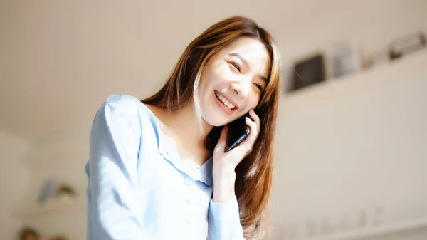 Happy young asian woman speaking on mobile phone at home, Female teen answering call on cellphone while sitting in couch. Asia girl talking on smartphone
