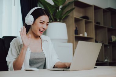 Evde bilgisayar bilgisayarı üzerinde çalışırken kulaklık takan Asyalı genç bir kadın. Evde çalışmak, video konferansı, video çağrısı, öğrenci öğrenme çevrimiçi sınıf