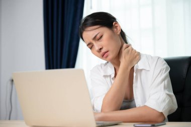 Ofis sendromu konsepti. Bilgisayarda uzun süre çalıştıktan sonra boyun ve omuz ağrısı çeken Asyalı genç bir kadın. Kaslarını gevşetmek için esniyor.